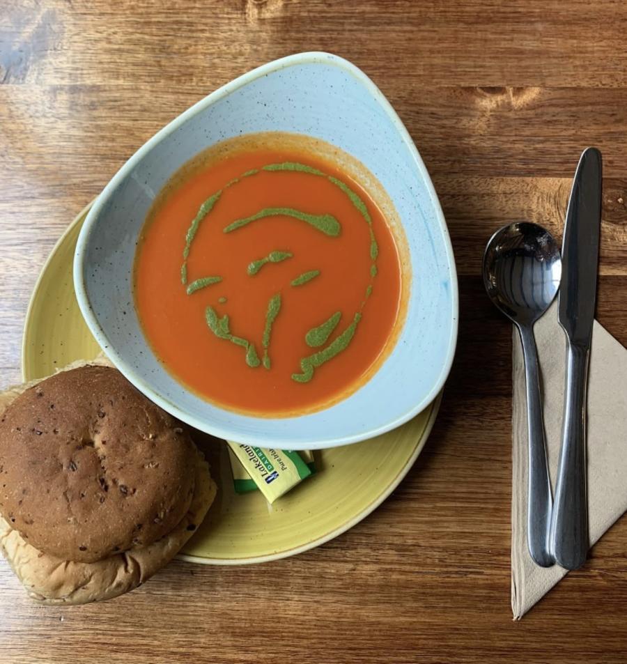 Book the Cotswolds - Wonderful Winchcombe Overhead view of a place setting on a wooden table featuring a bowl of smooth tomato soup drizzled with green herb oil. The bowl sits on a light green plate, accompanied by a bread roll. To the right are a neatly placed knife and fork on a folded napkin, along with a small condiment container. The scene is warmly lit, highlighting the textures of the wood and the vibrant color of the soup.