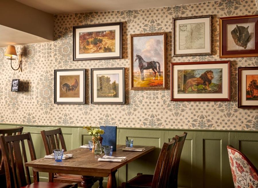 Book the Cotswolds - Wonderful Winchcombe Inside the cosy restaurant at The Lion Inn. A wooden table set for four, surrounded by dark wooden chairs. The wall behind features patterned wallpaper and is decorated with a gallery of framed artwork, many depicting horses and animals in various styles and sizes. A small vase of yellow flowers sits on the table alongside menus, plates, and cutlery. Warm, ambient lighting creates an inviting, traditional atmosphere.